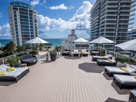 A stylish rooftop lounge with multiple sunbeds and umbrellas, overlooking the ocean and tall apartment buildings under a bright blue sky.