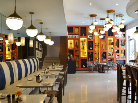 A stylish restaurant interior with a long booth seating on the left, bright pendant lights, and a red display wall with shelves and chairs on the right.