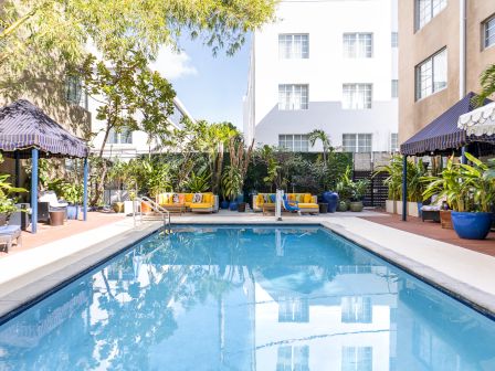 A sunlit hotel pool area with clear blue water, lounge chairs, shaded cabanas, potted plants, and a modern building backdrop.