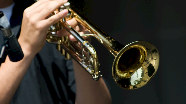 A person plays a shiny gold trumpet on stage, holding it up toward the camera with fingers pressing the valves.